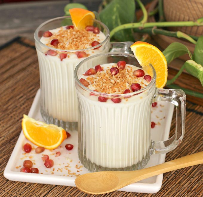 White chocolate coconut mousse topped with toasted coconut and pomegranate seeds in clear glass cups with an orange wedge