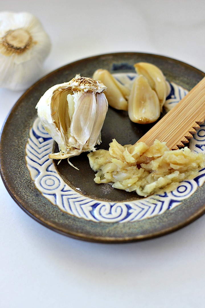 Brown plate with partial bulb or roasted garlic, 3 whole roasted garlic cloves and some mashed roasted garlic with t wooden knife.