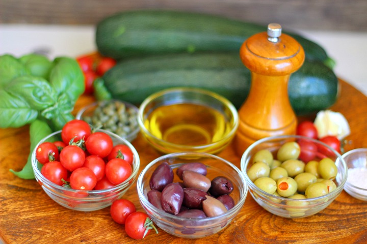 Bowls of cherry tomatoes, kalamata olives, green olives, olive oil, capers, salt, pepper grinder, 2 zucchini, garlic and fresh basil