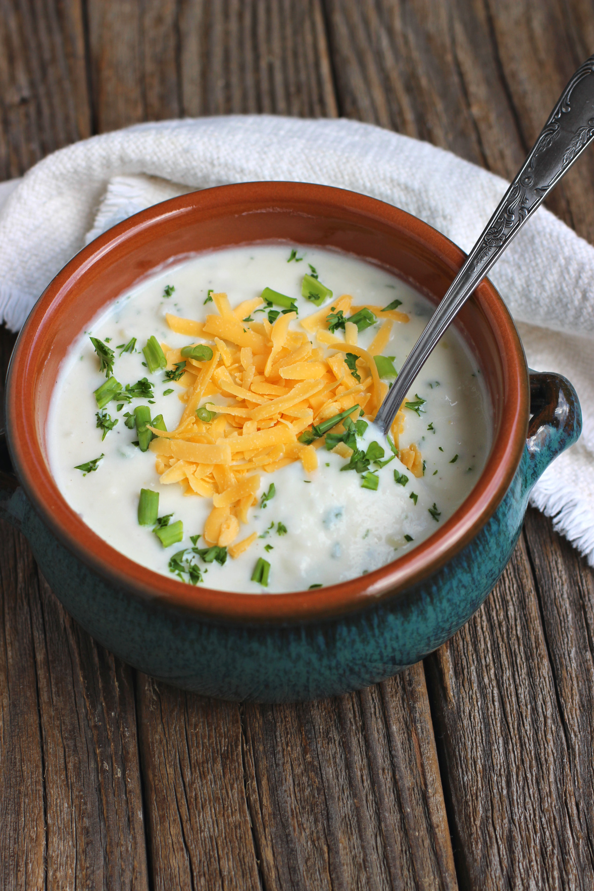 blue crock of potato soup with spoon and garnished with shredded cheese and parsley on rustic surface