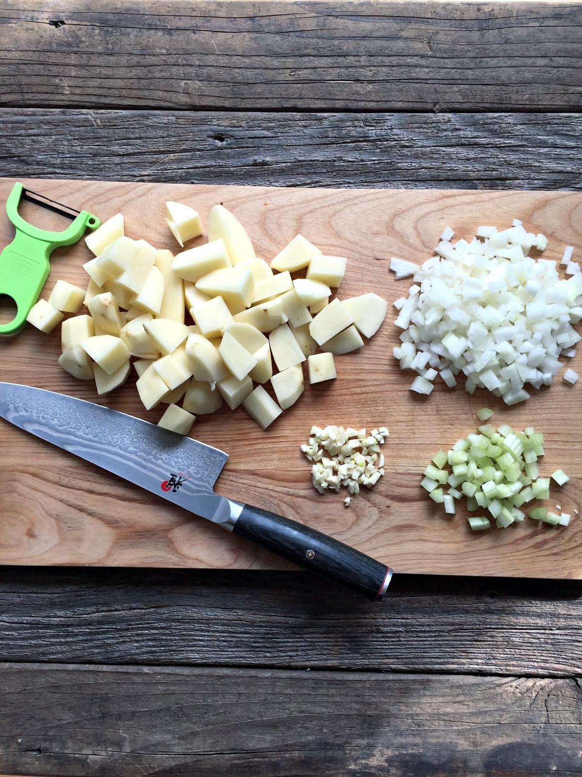 cutting board with knife and peeler, chopped potatoes, onions, celery and garlic