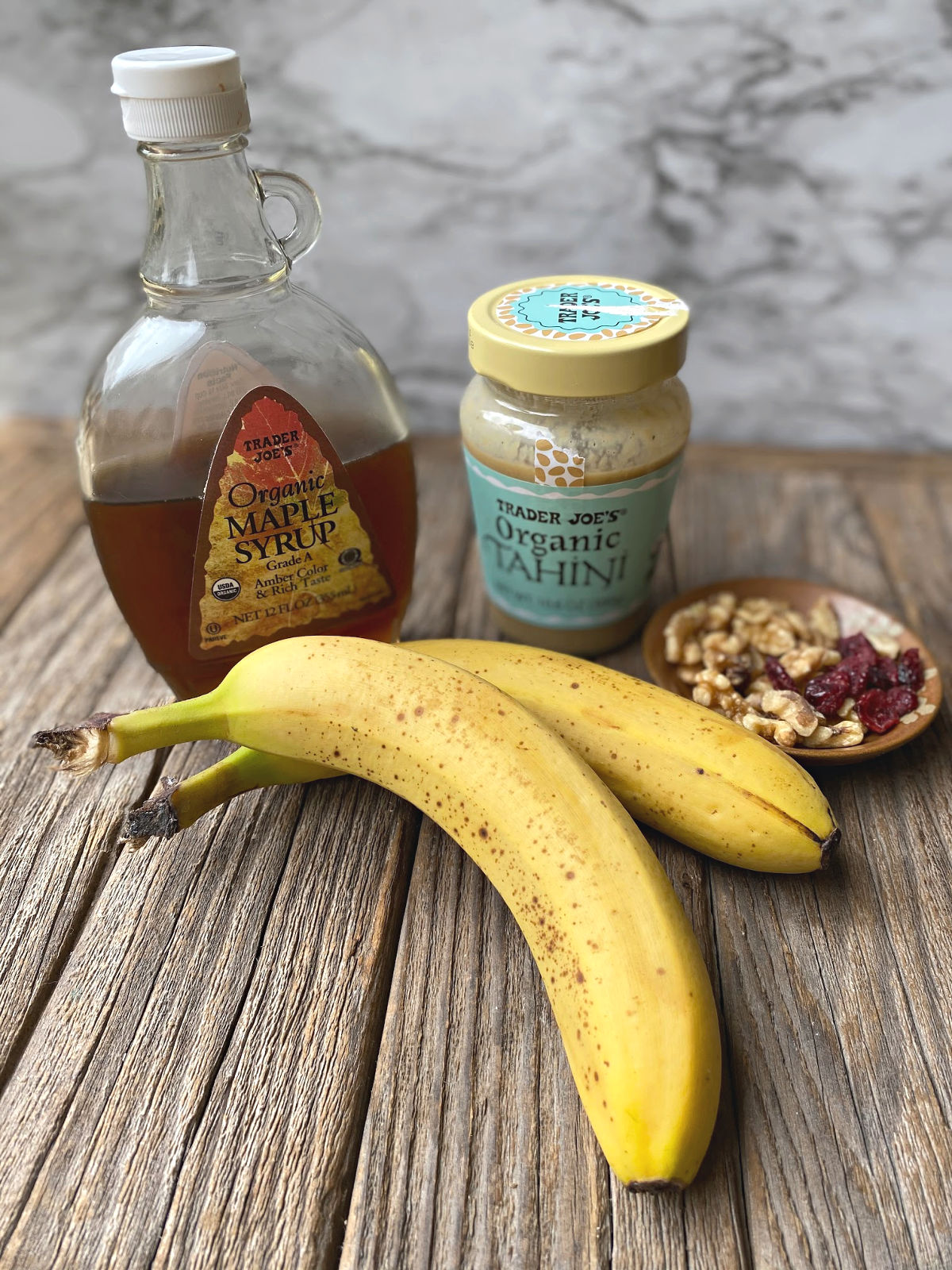 rustic surface with 2 bananas, maple syrup, tahini and small bowl of walnuts and dried cranberries